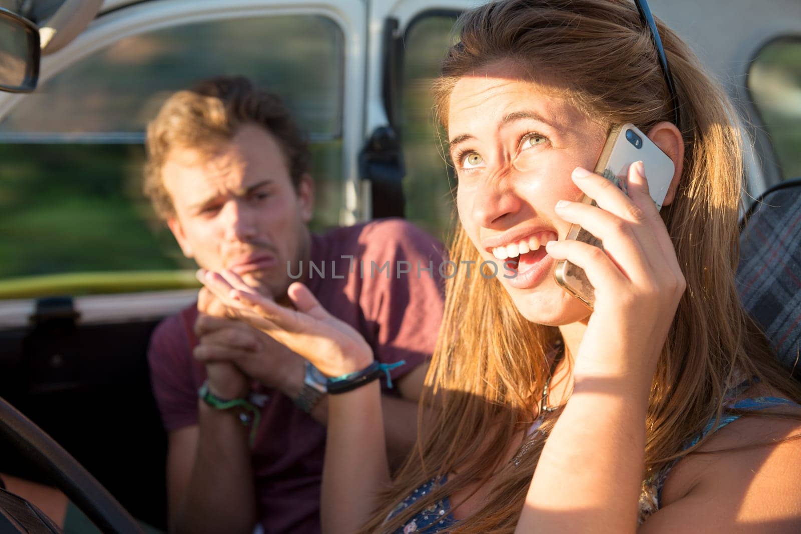 Couple in a car at sunset, with girl driving and speaking at the phone, boy scared and praying.