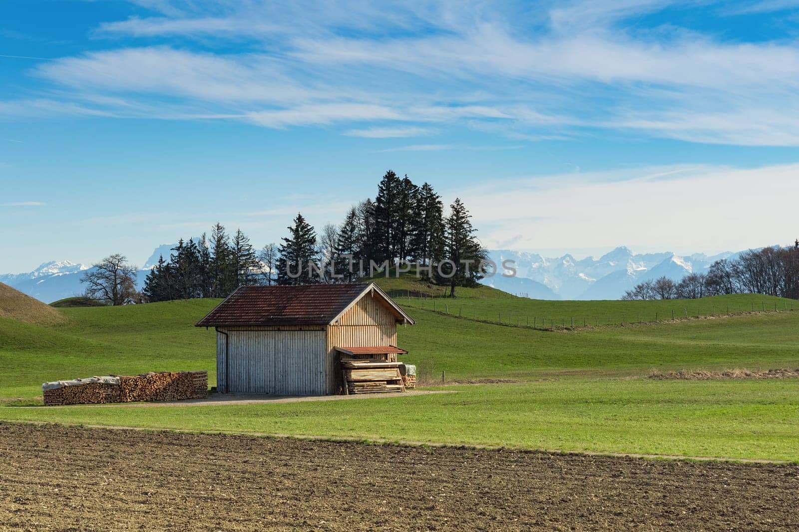 Alpine mountains spring landscape with sunny green grassland field and wooden shed