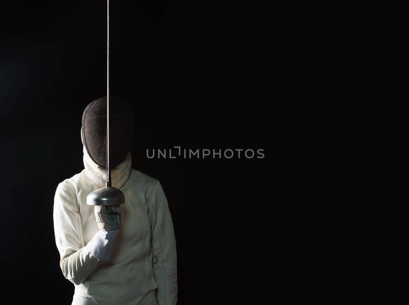 The portrait of woman wearing white fencing costume and black fencing mask with the rapier on black background