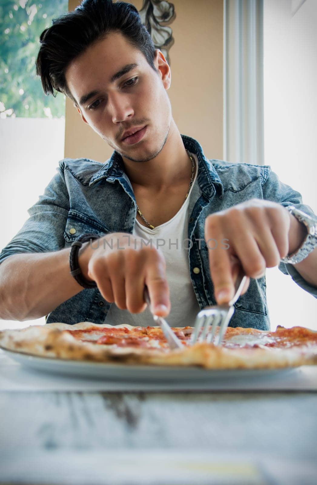 Low angle view of a young man eating an Italian pizza cutting it with a knife and fork