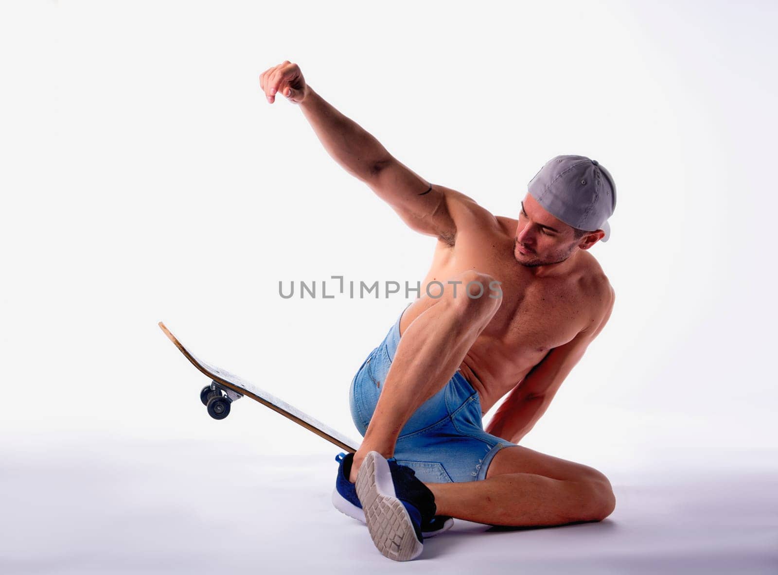 Shirtless Man Sitting on Skateboard in Studio with White Background Posing as if in Mid Trick