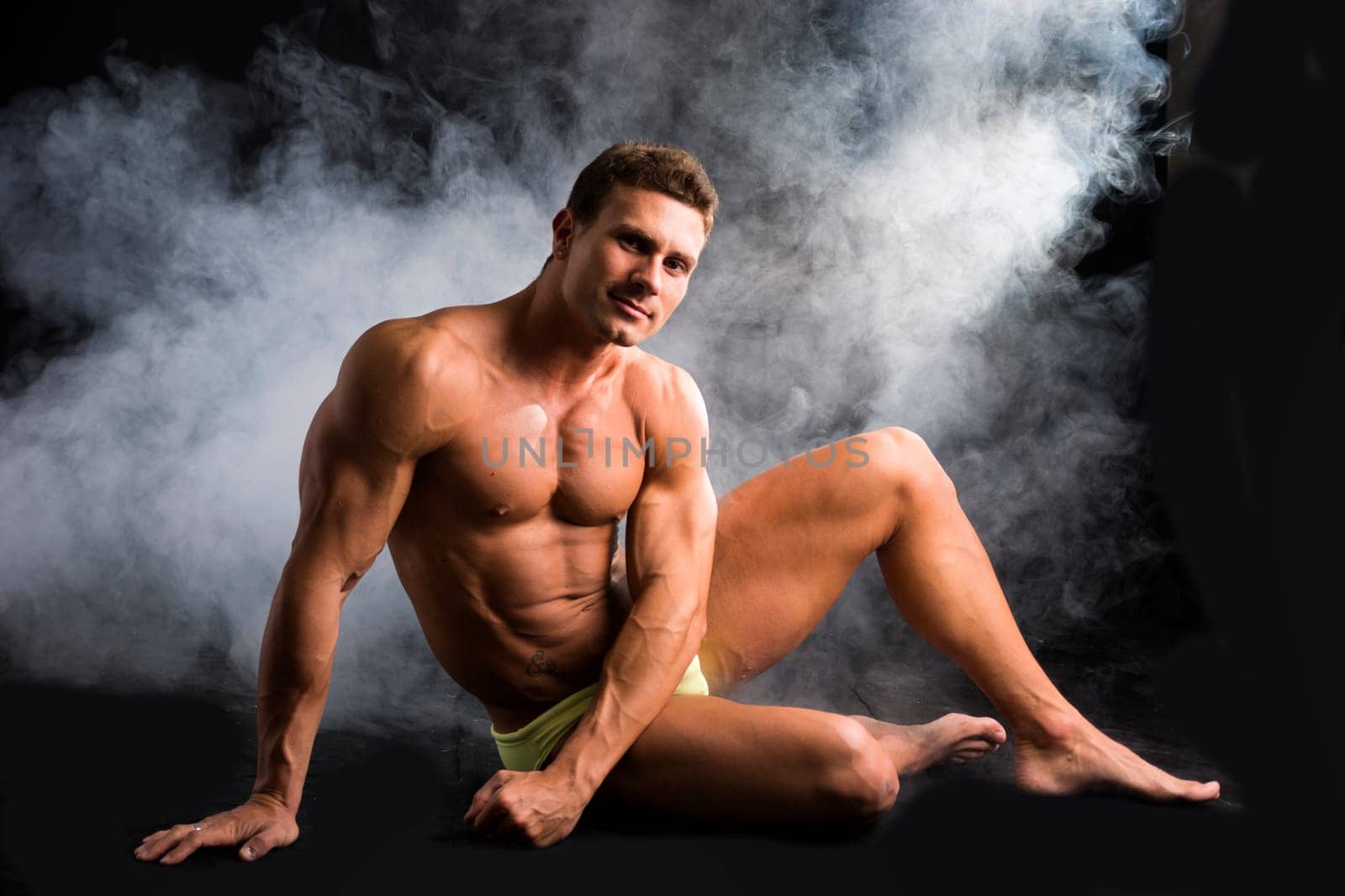 Attractive shirtless muscular man sitting on the floor in bathing suit on dark background