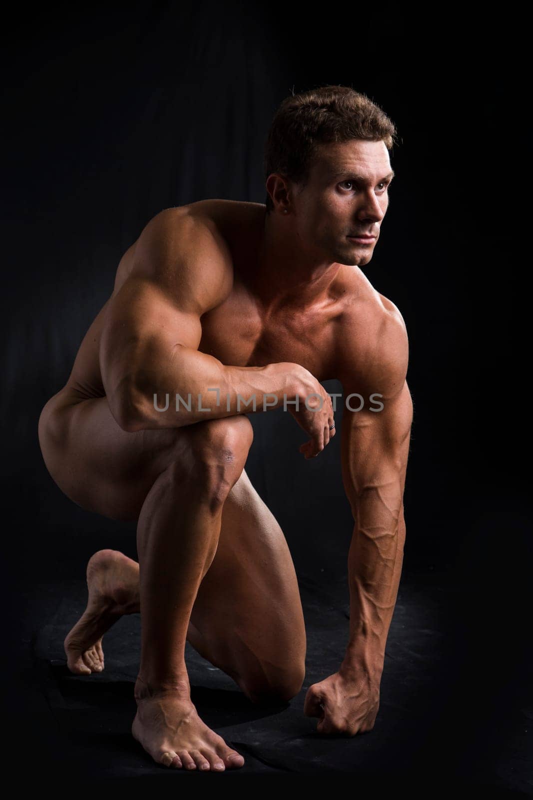 Totally Nude Young Muscle Man Crouching on one Knee in Studio with Black Background