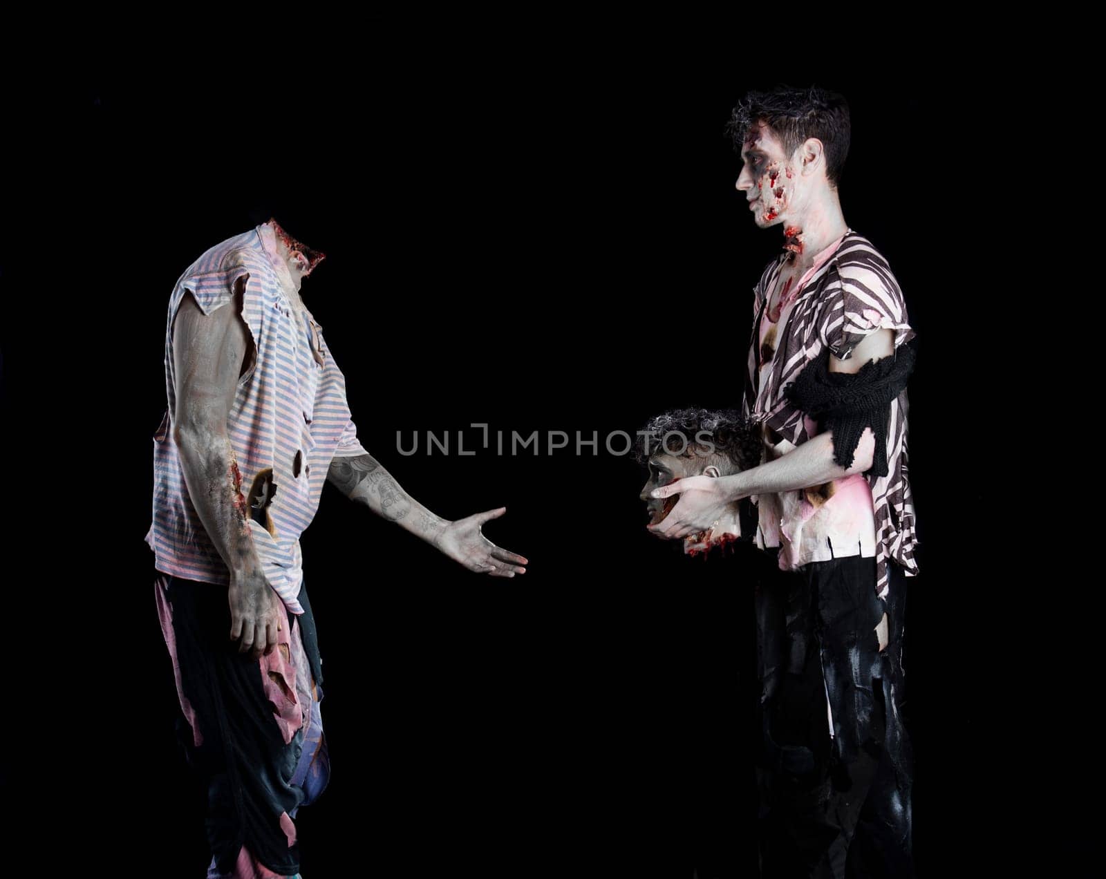 Two male zombies, one giving the head to the decapitated one's body, standing on black background