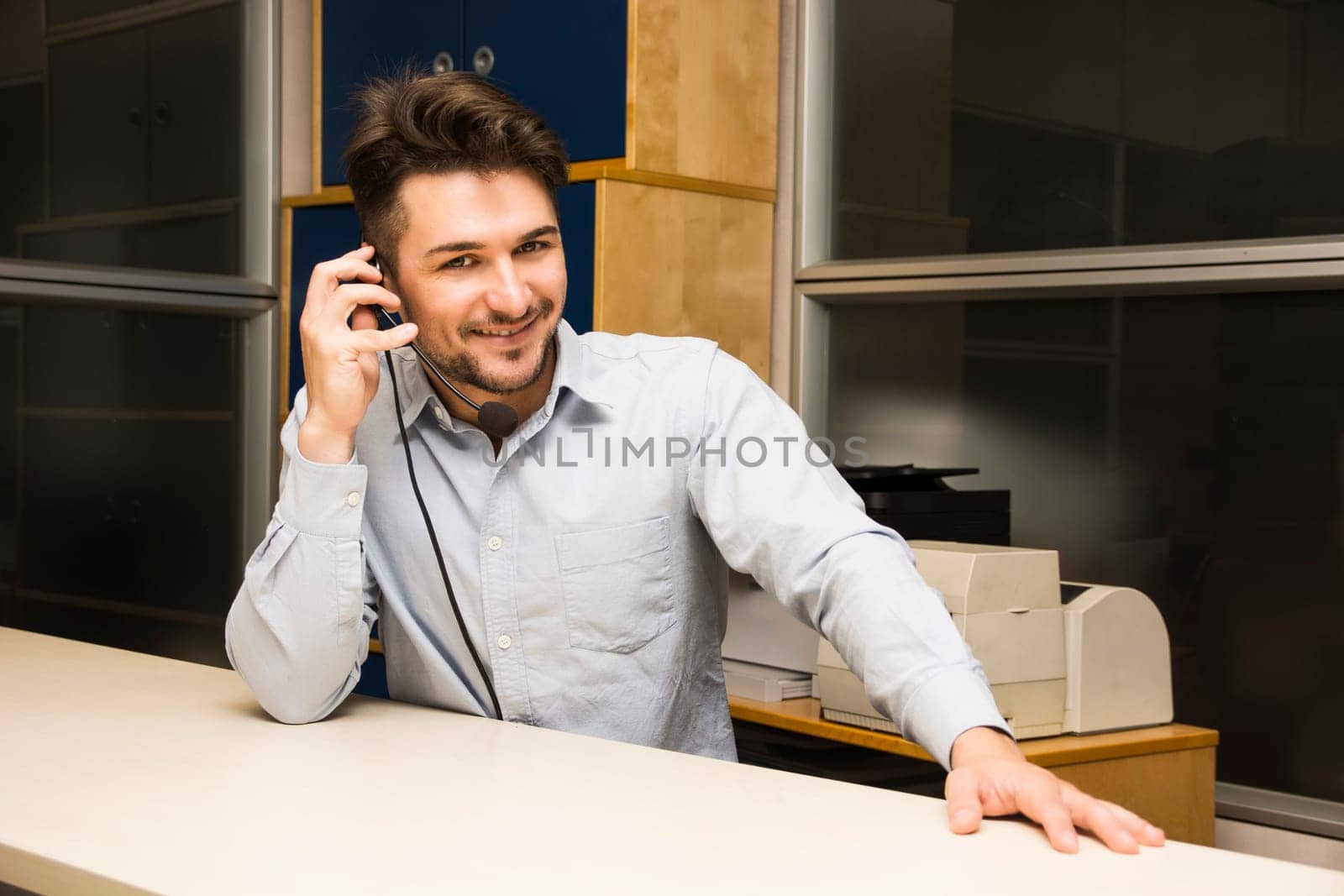 Businessman or customer (support) service man taking a call on his headset phone as he sits at a table in the office looking up to smile at the camera as he listens to the conversation
