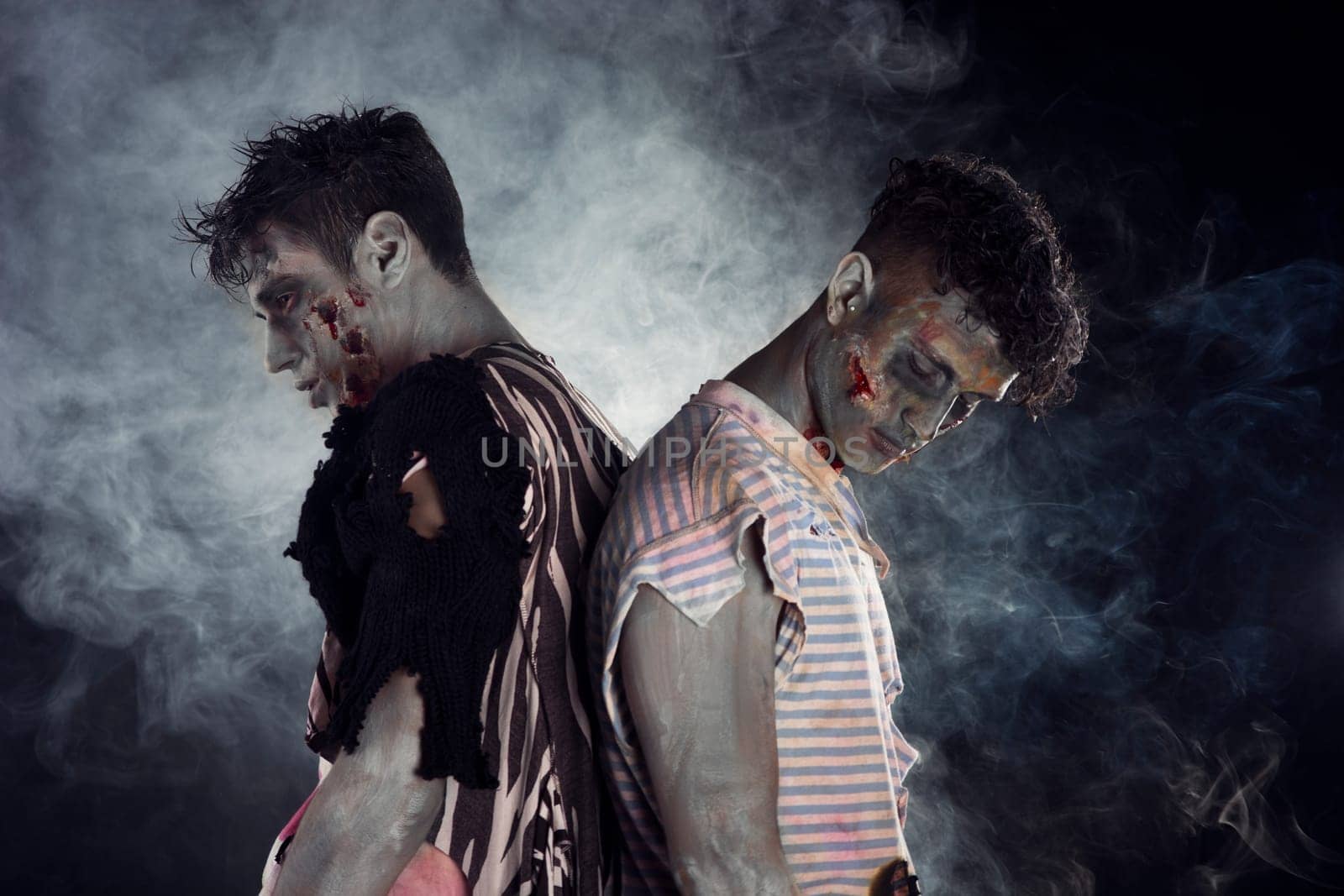 Two male zombies back to back on black smoky background, looking down and away