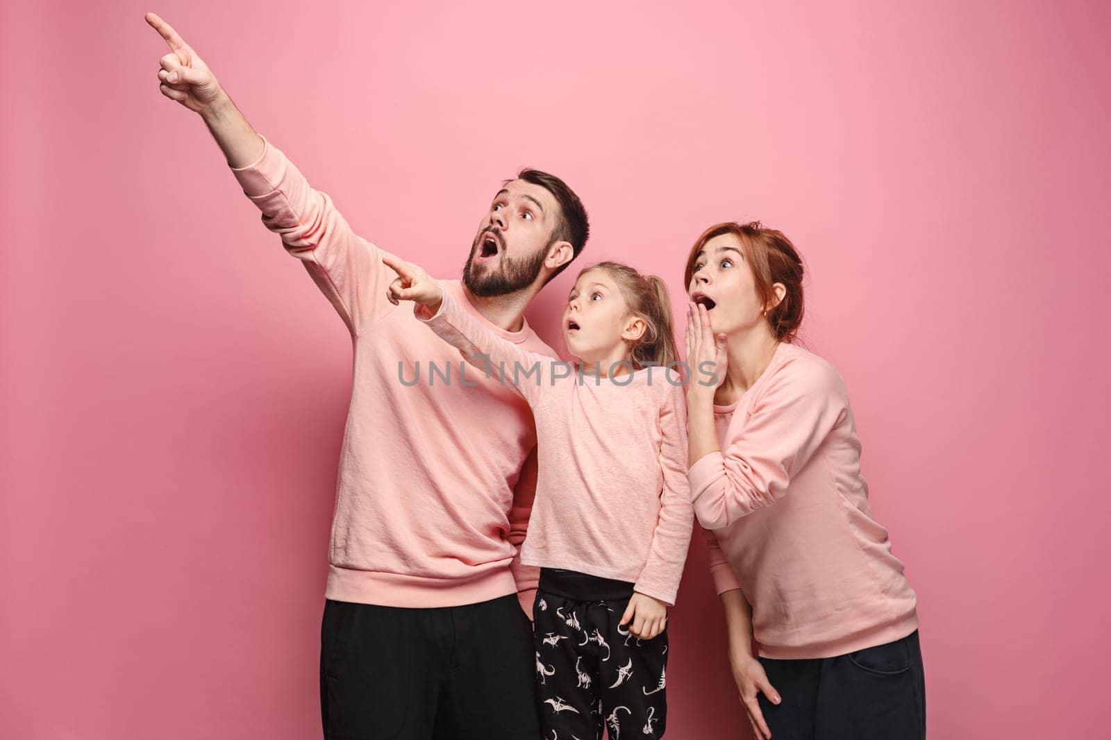 Surprised young family pointing up by fingers on pink studio background