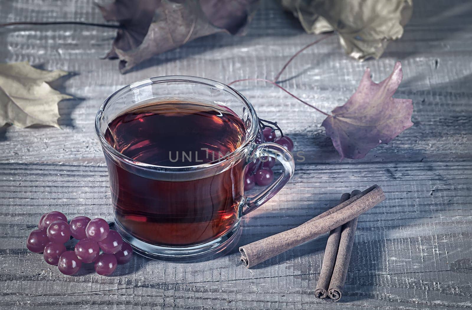 Still life with transparent and fragrant Cup of tea with viburnum and cinnamon on wooden table. Dry autumn maple leaves in the background. Toned photo.