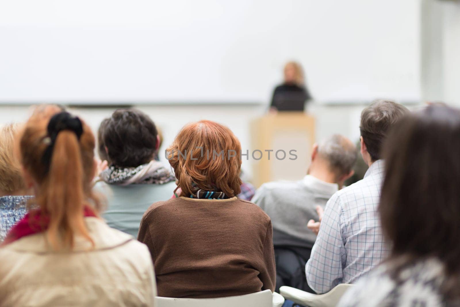 Business and entrepreneurship symposium. Female speaker giving a talk at business meeting. Audience in conference hall. Rear view of unrecognized participant in audience. Copy space on whitescreen.
