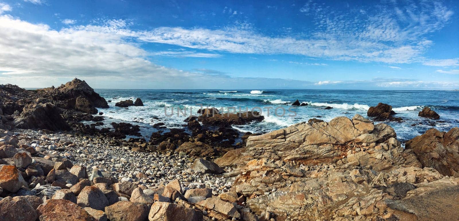Panorama of Pacific ocean cost in California, USA. Rocky beach in foreground and blue cloudy sky in background.