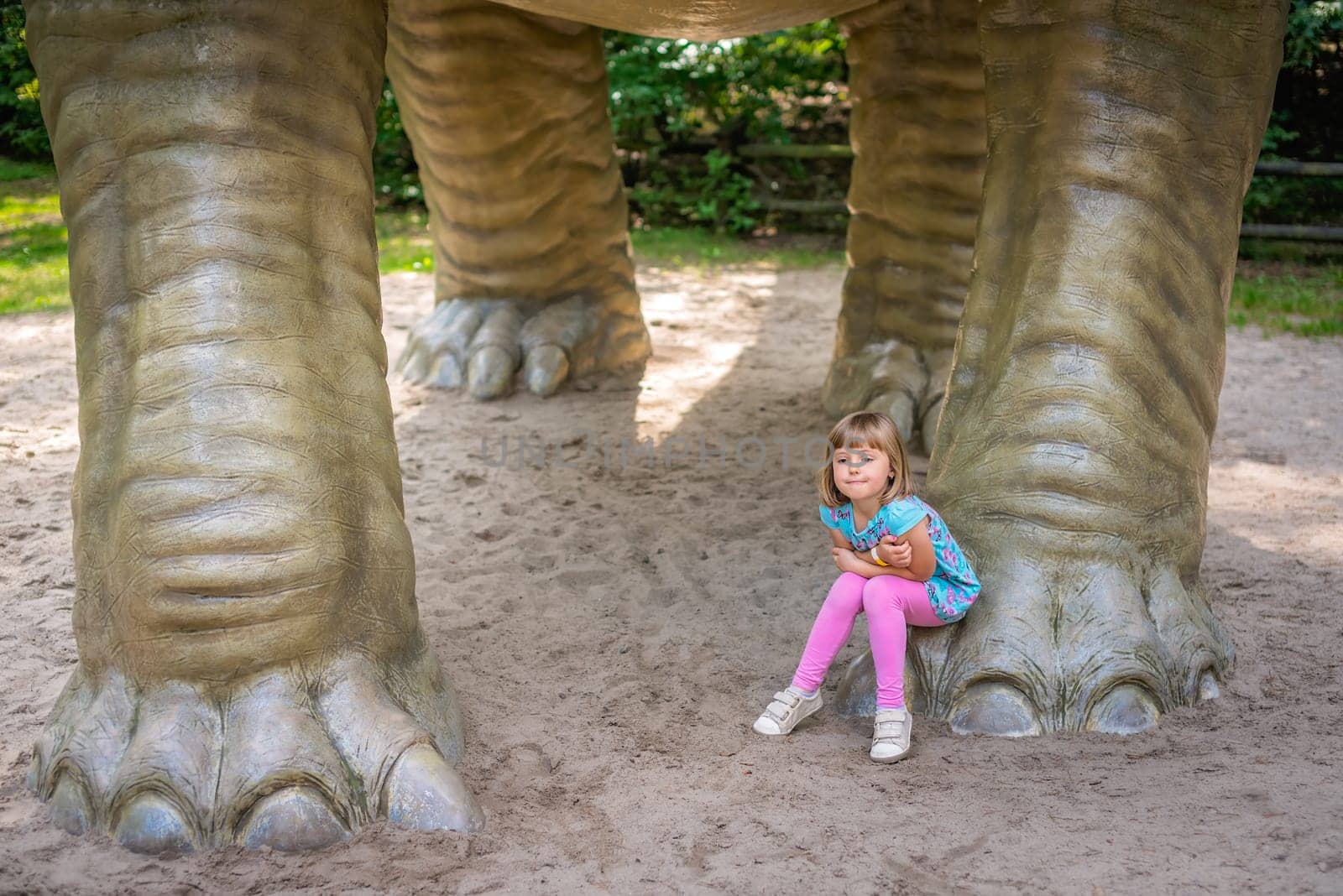 Little girl sitting under huge life sized Diplodocus dinosaur sculpture in an amusement park in summer