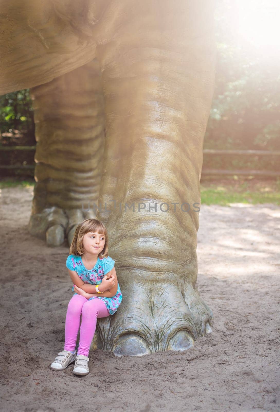 Little girl sitting under huge life sized Diplodocus dinosaur sculpture in an amusement park in summer