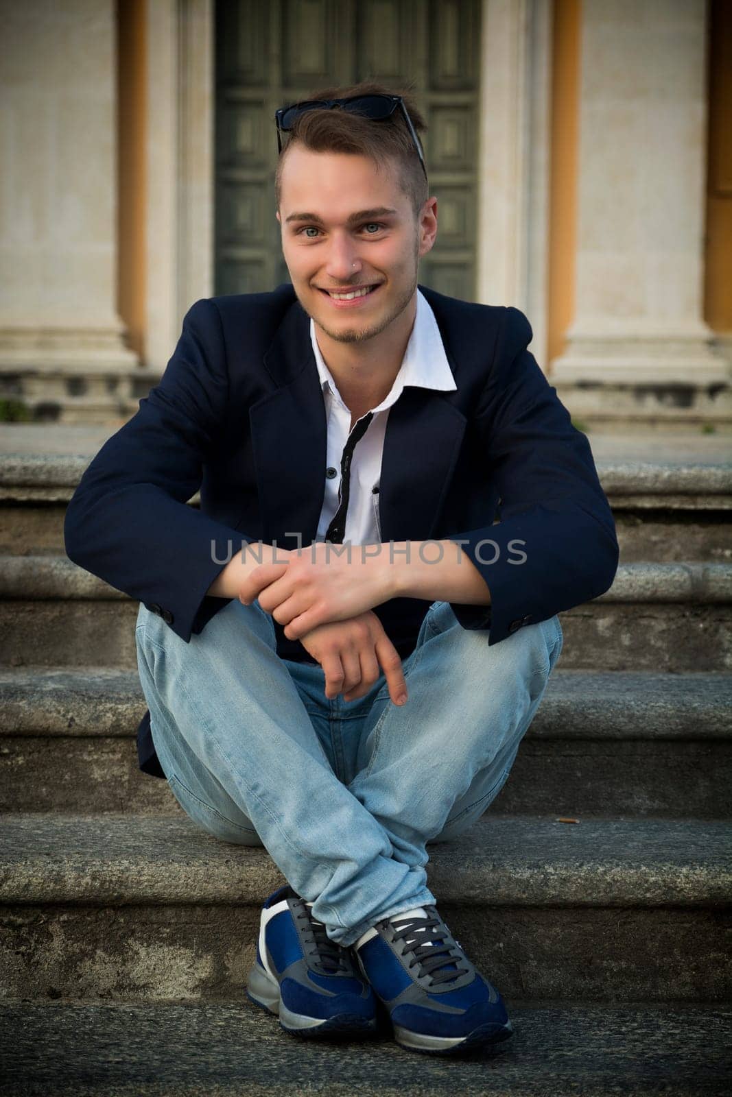 Smiling blond young man sitting on stone stair steps outside, looking at camera