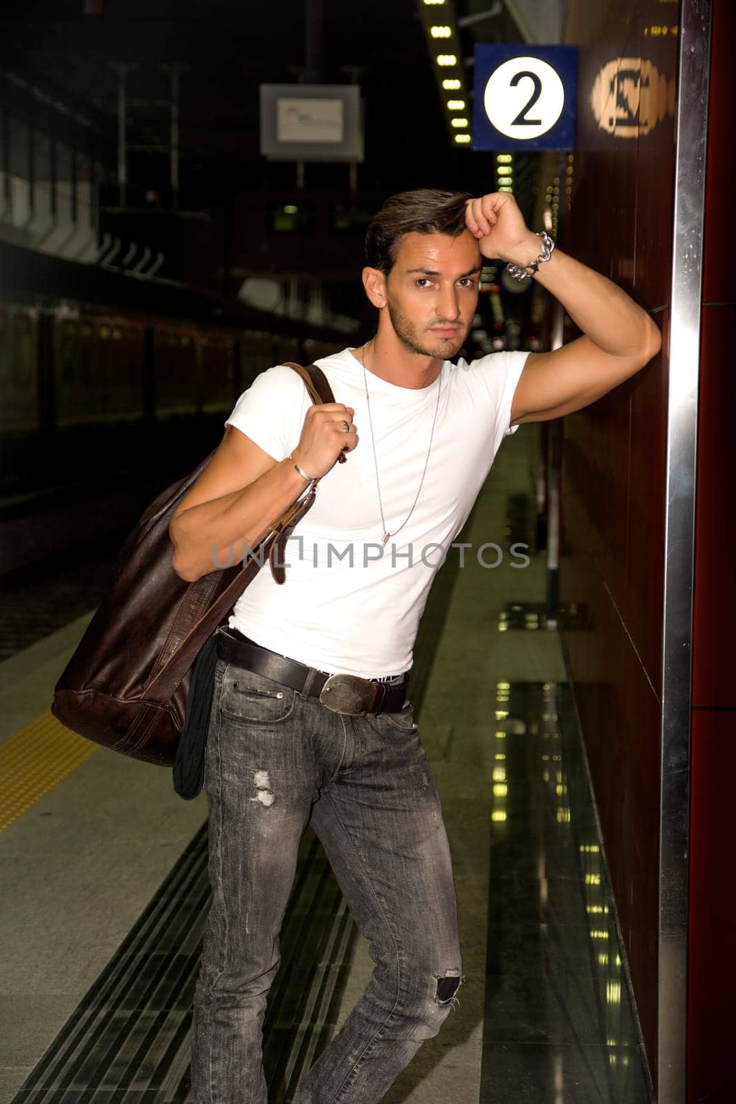 Handsome young man standing against wall in train or subway station, looking at camera