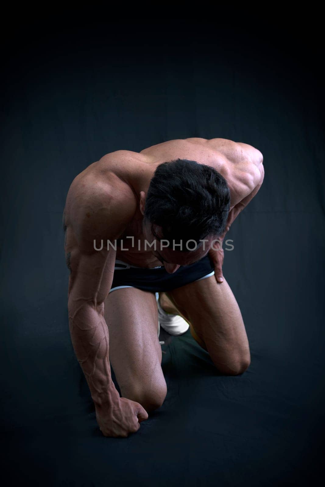 Muscular male bodybuilder kneeling with head down on dark background
