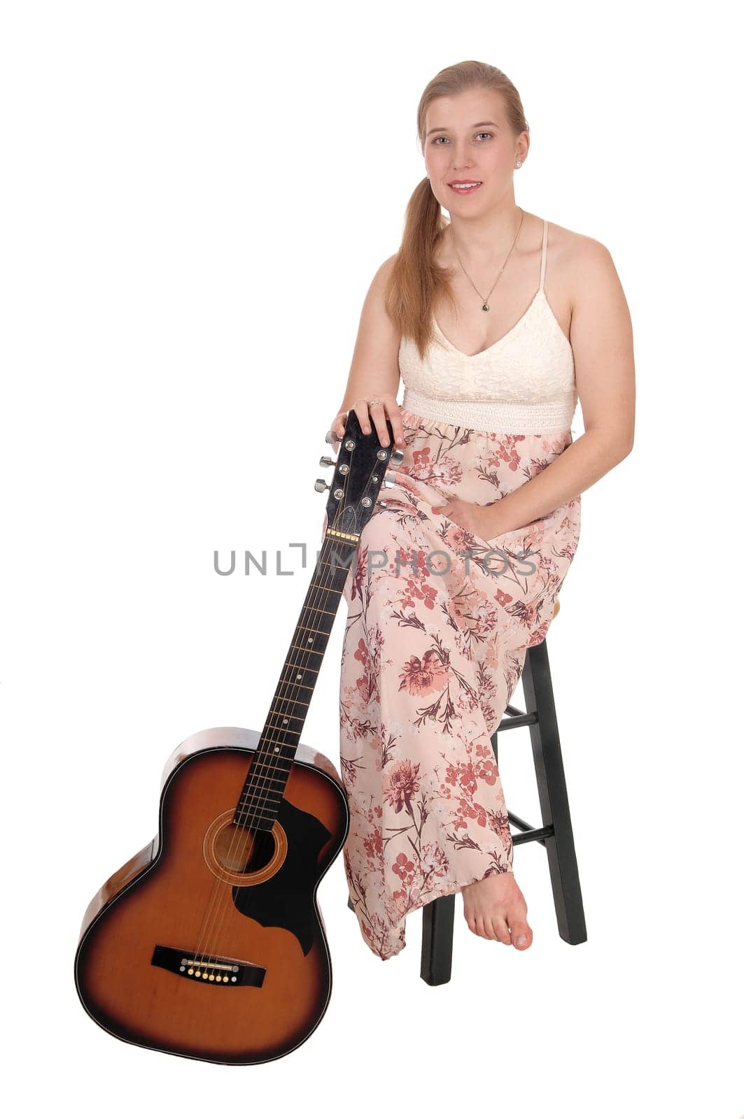 A beautiful blond woman in a long dress sitting on a bar chair with.her guitar on her side, isolated for white background.