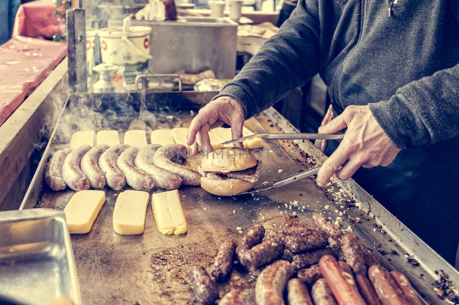 Hands of the cook preparing a hot sandwich on a cooking plate where sausages and frankfurters are cooking. Street food.