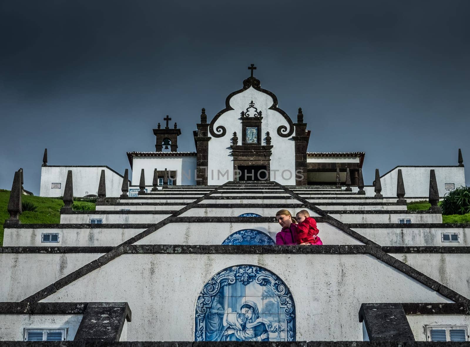 Mother carrying her son while walking up the stairs leading to the entrance of the Our Lady of Peace ( Nossa Senhora da Paz ) chapel above the city of Vila Franca do Campo in Sao Miguel, Azores, Portugal