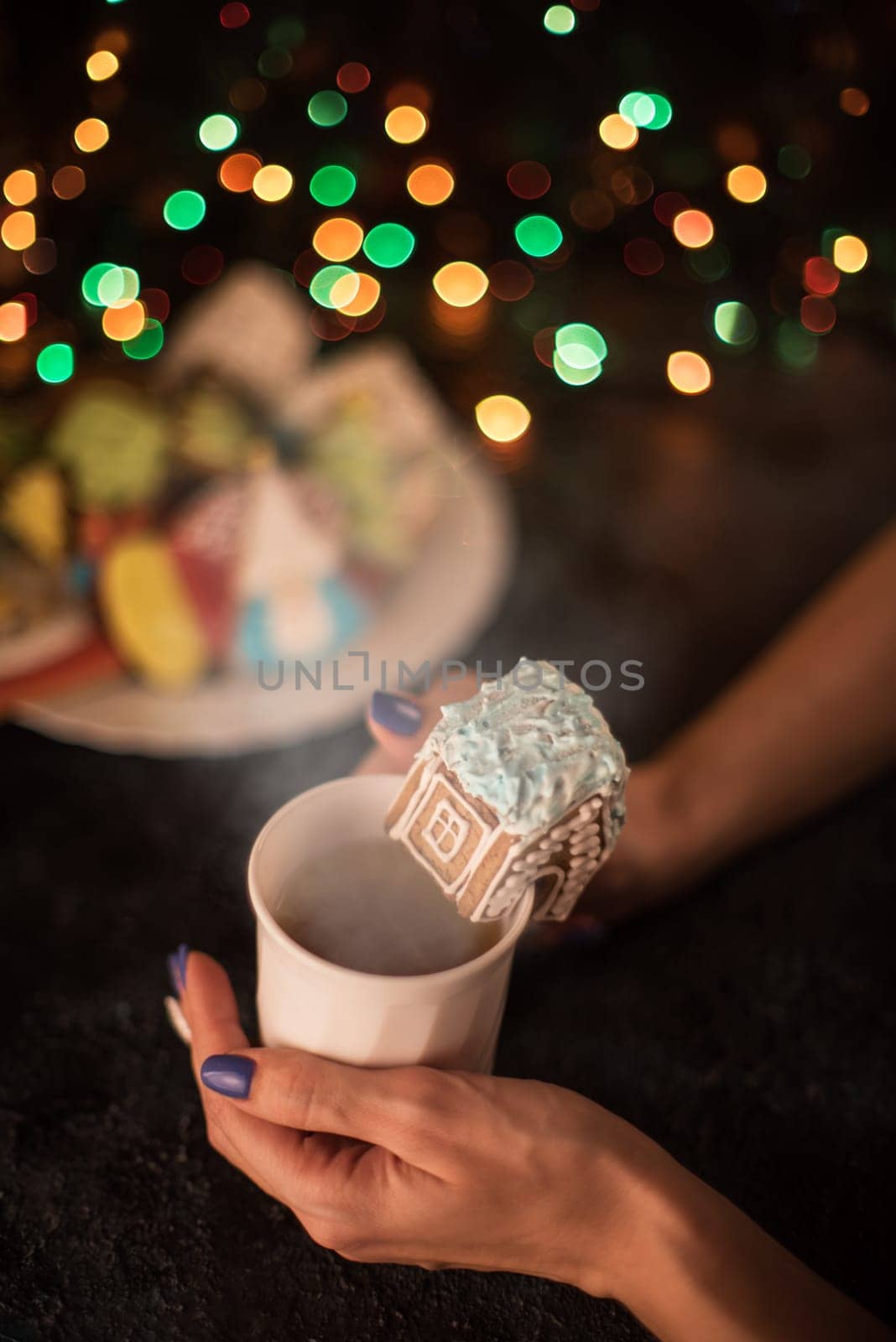 Female hands with cup of tea and christmas cookies house on dark color bokeh lights background