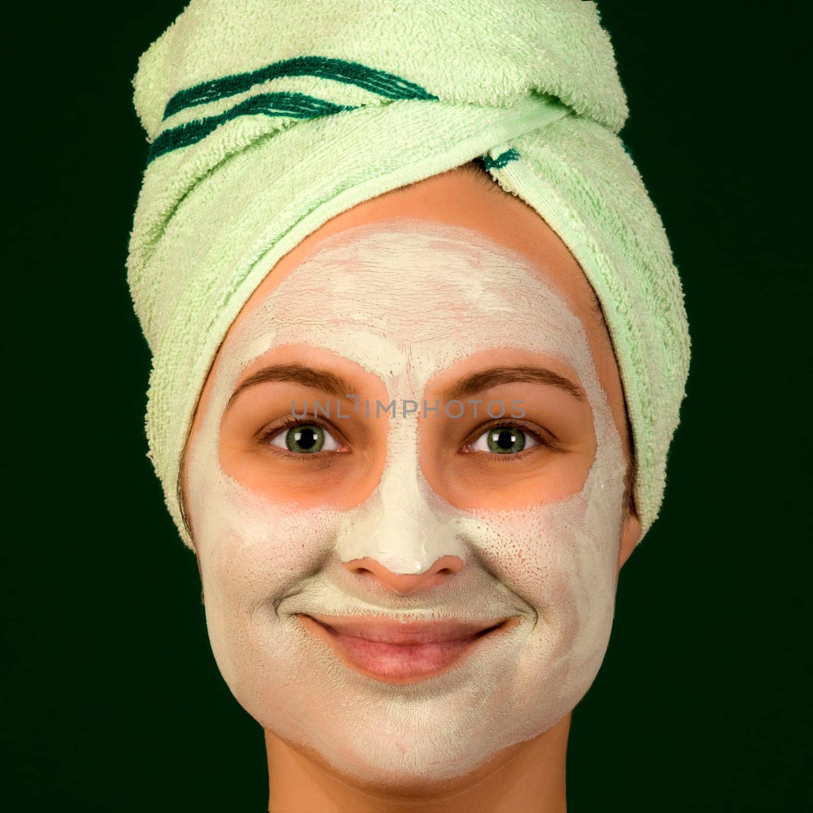 Portrait of woman with face mask and towel on dark green background. Cosmetology concept.