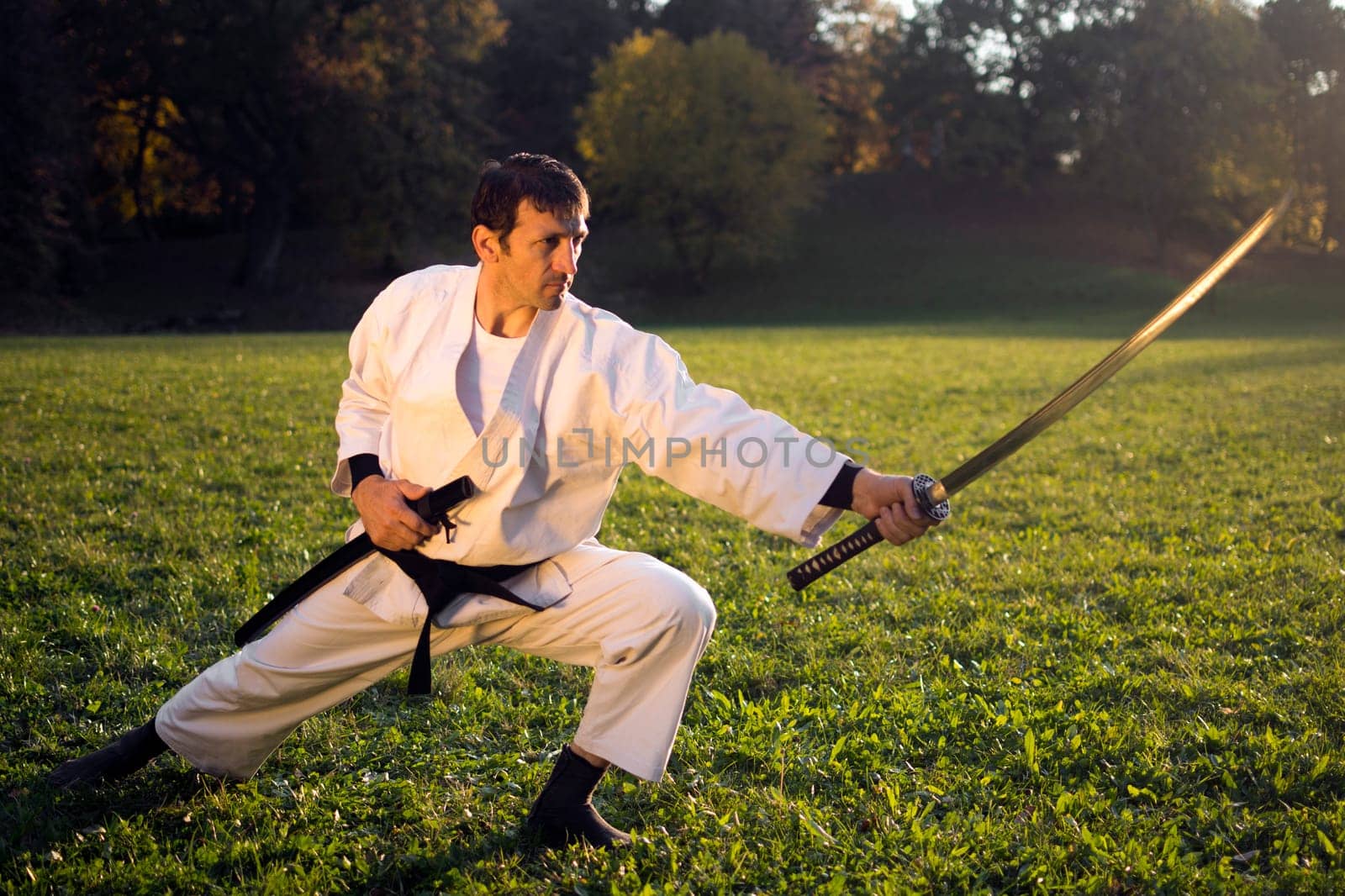 Ninja in white kimono with black belt is practicing with sword outdoors in the nature.