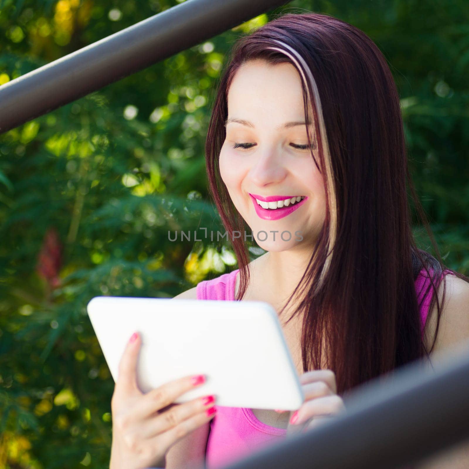 Smiling woman is using her white digital tablet outdoors in nature on green background.