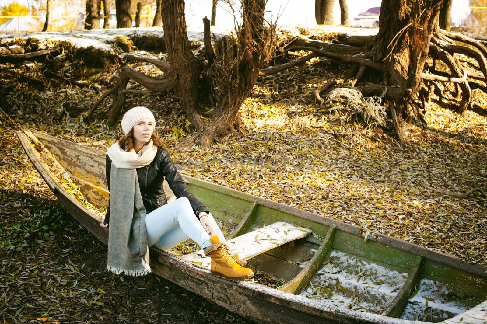 Slim girl in blue jeans, brown jacket and boots, sat on the upturned boat, on the shore, yellow autumn leaves bright sunny day