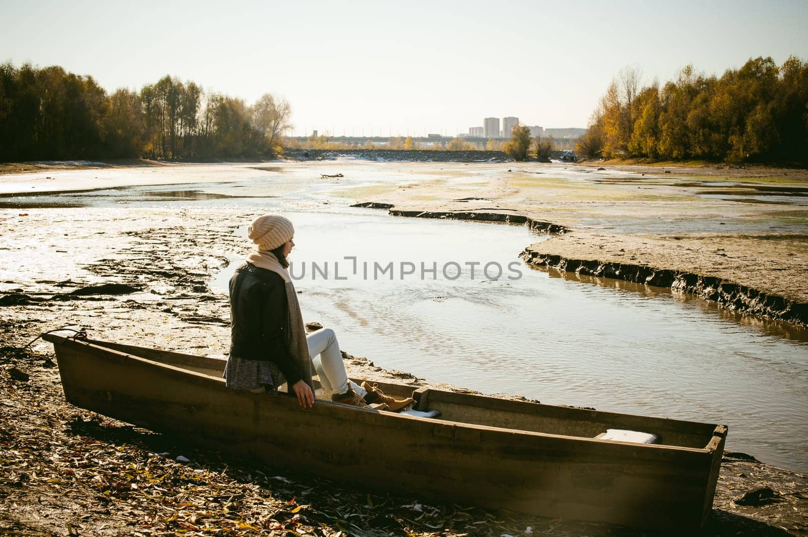 Slim girl in blue jeans, brown jacket and boots, sat on the upturned boat, on the shore, yellow autumn leaves bright sunny day