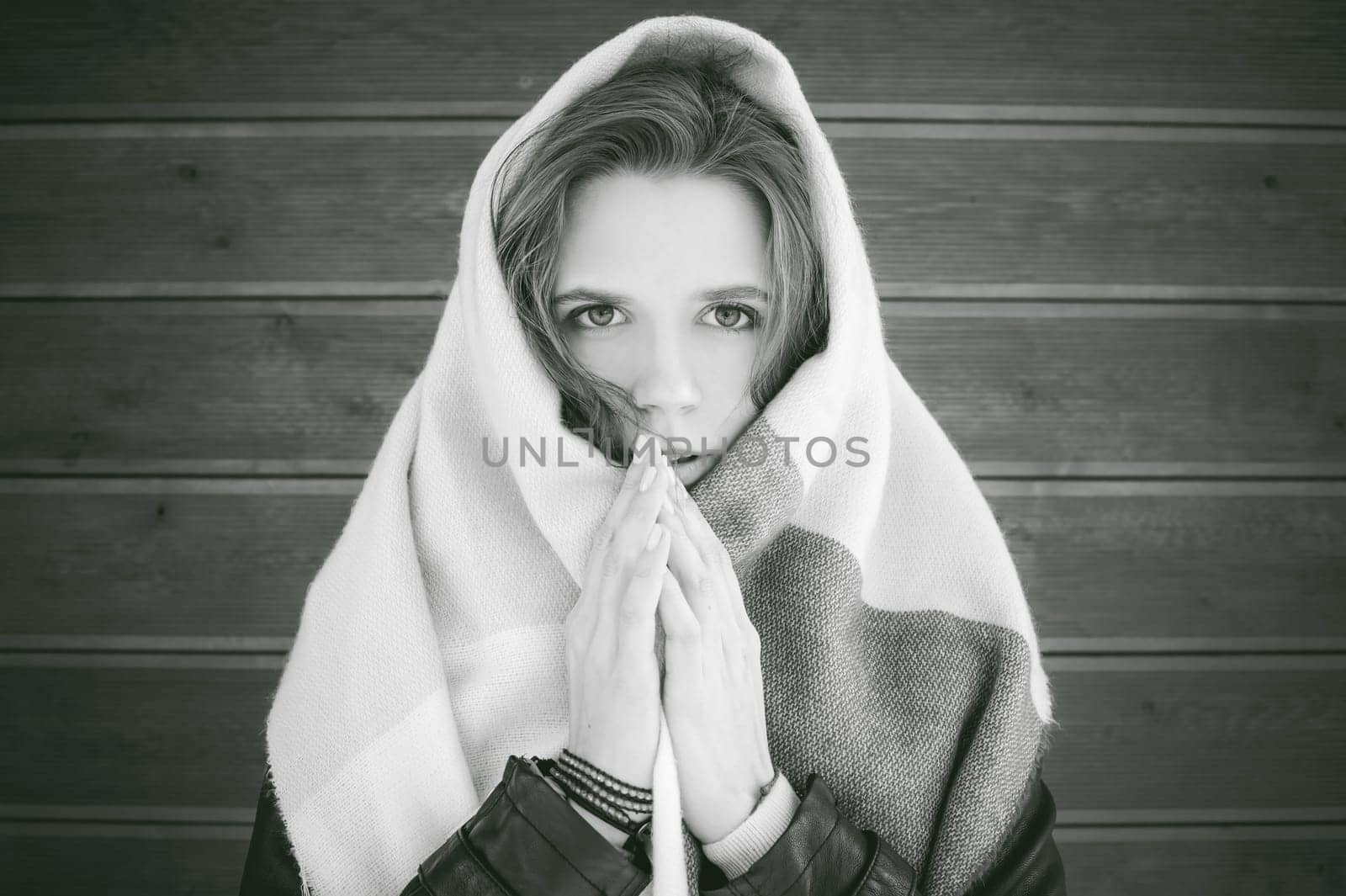 Portrait of young woman walking outdoors on warm autumn afternoon. On the background of a wooden wall, wrapped in a scarf with his head, holding his hands in front of his face