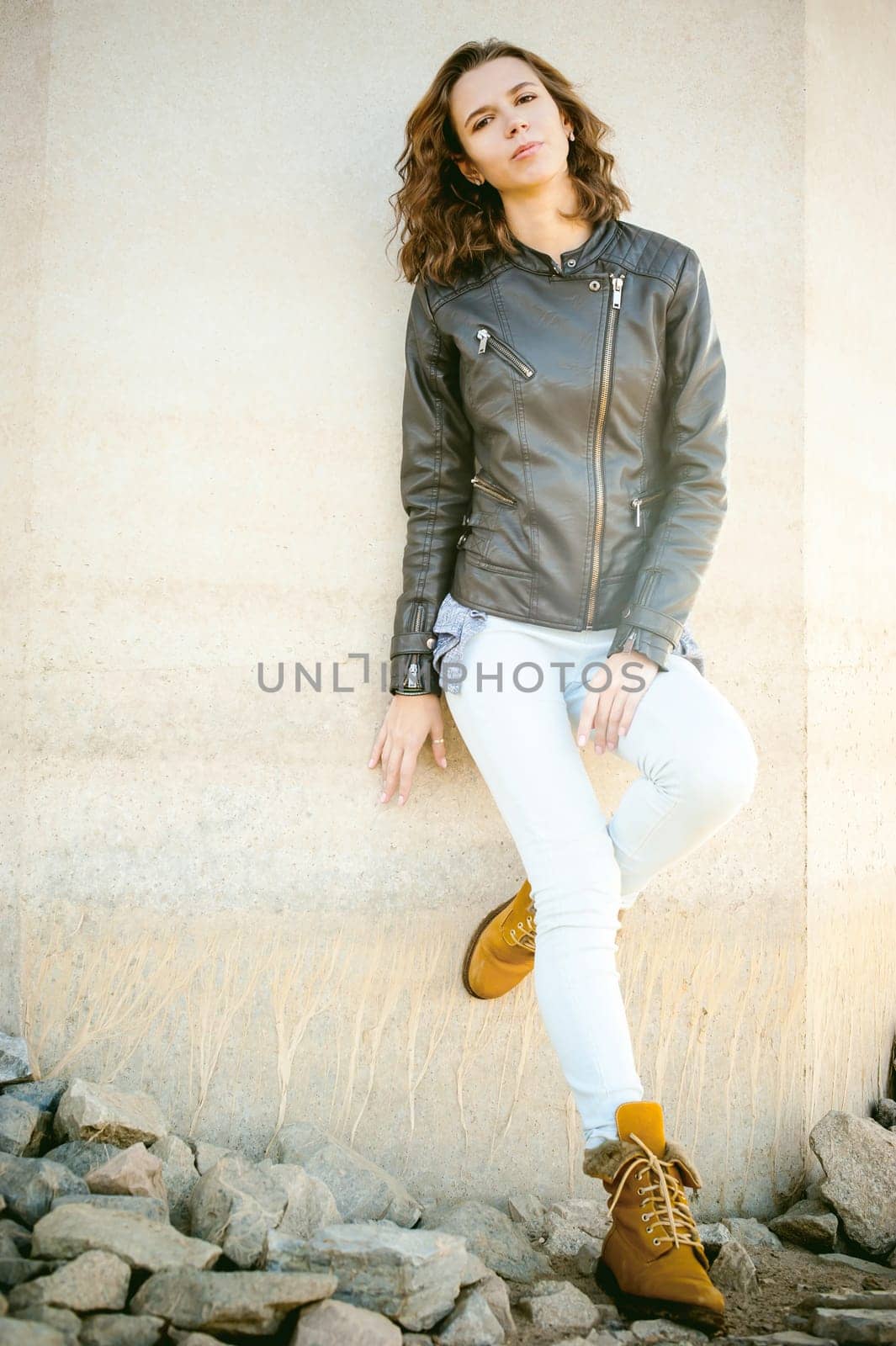 beautiful legs of the young girl in blue jeans, a leather jacket and brown boots. woman standing his back against the concrete wall, the stones beneath her feet. lit by beautiful warm autumn sunshine