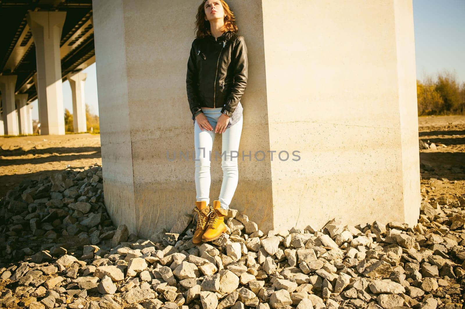 beautiful legs of the young girl in blue jeans, a leather jacket and brown boots. woman standing his back against the concrete wall, the stones beneath her feet. lit by beautiful warm autumn sunshine