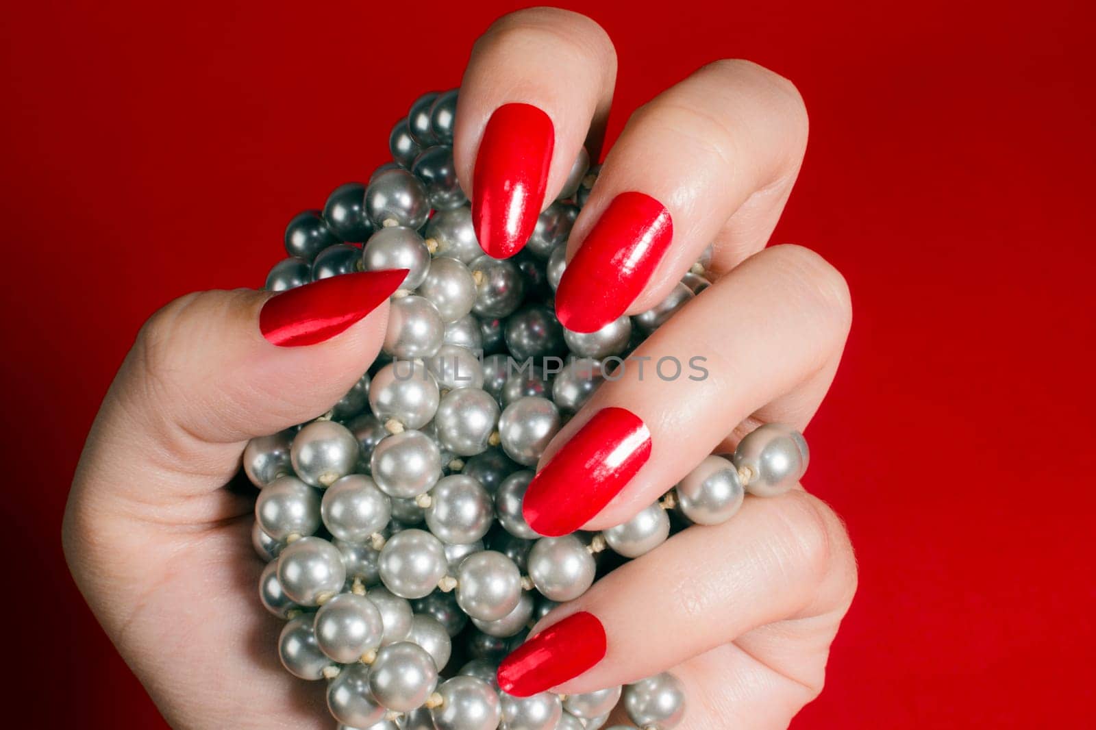 Female hand with red nails is holding pearls on red background. Manicure concept.