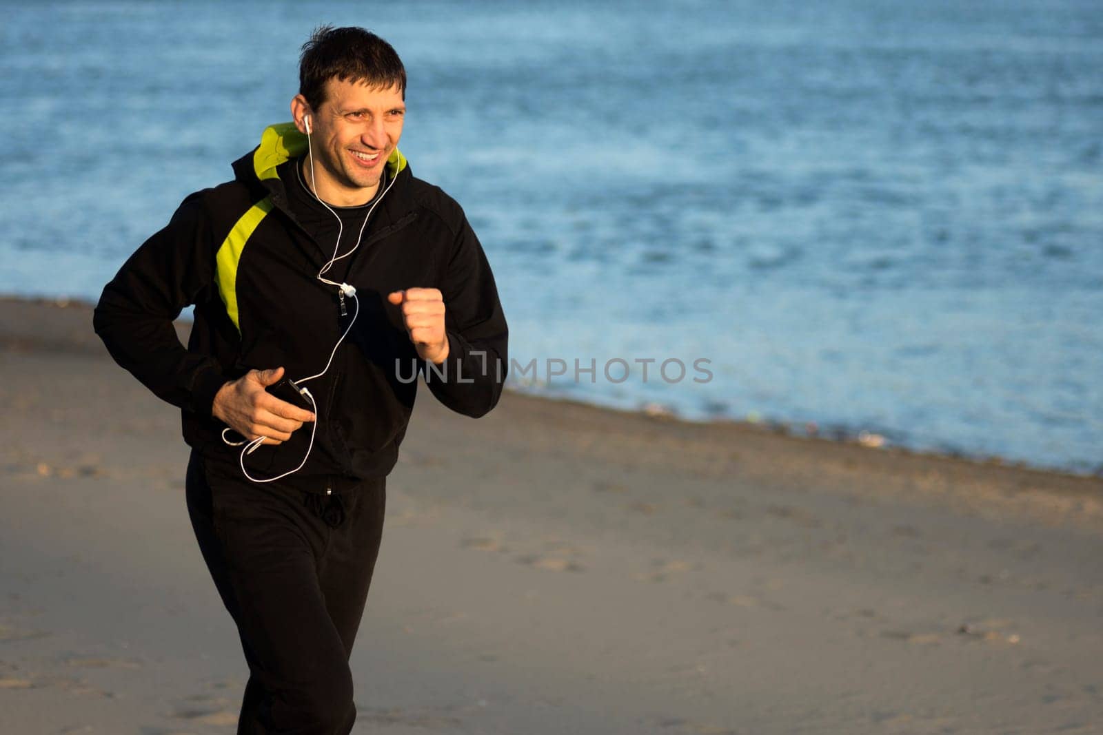 Smiling man with earphones is jogging along the coast.