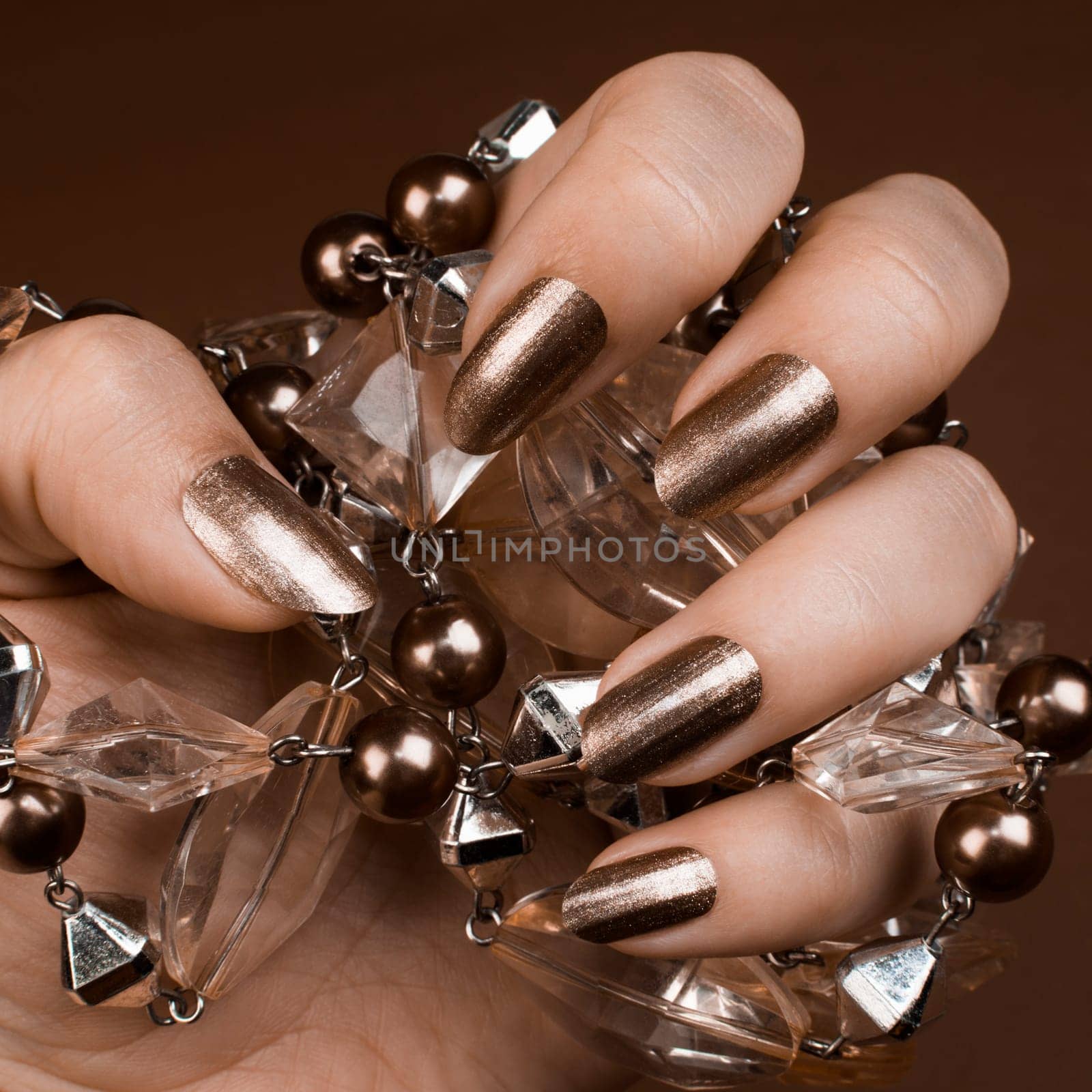 Female hand with shiny brown nails holds brown jewel on brown background.