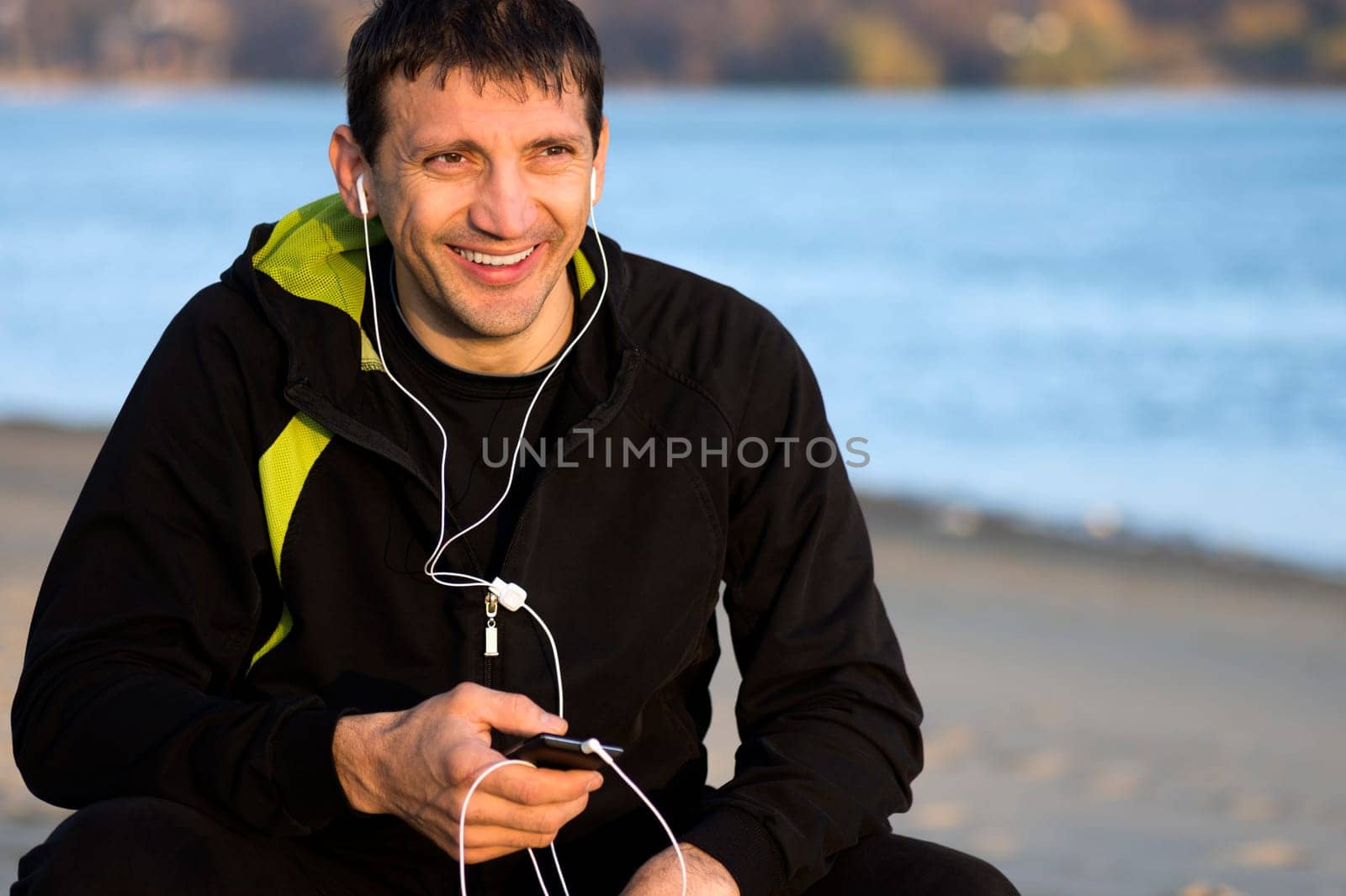 Smiling man with earphones is sitting outdoors along the riverside. Lifestyle concept.