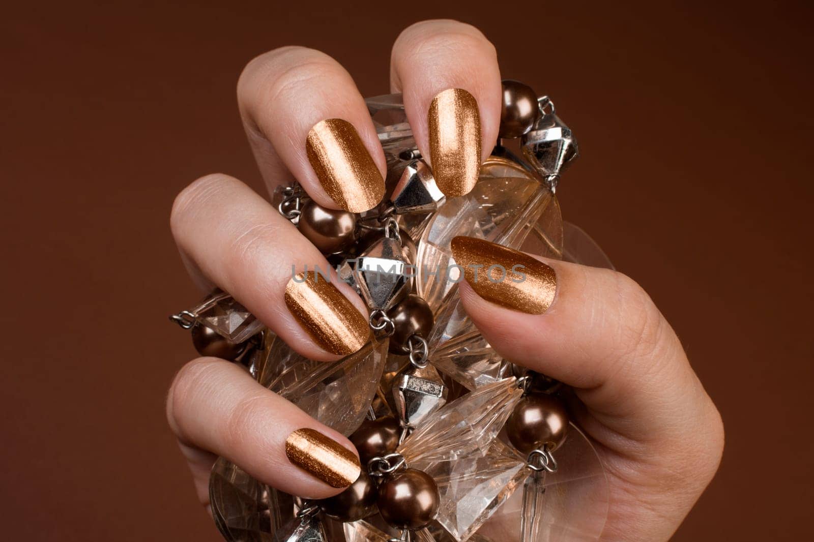Female hand with shiny golden nails is holding brown jewel on brown background.
