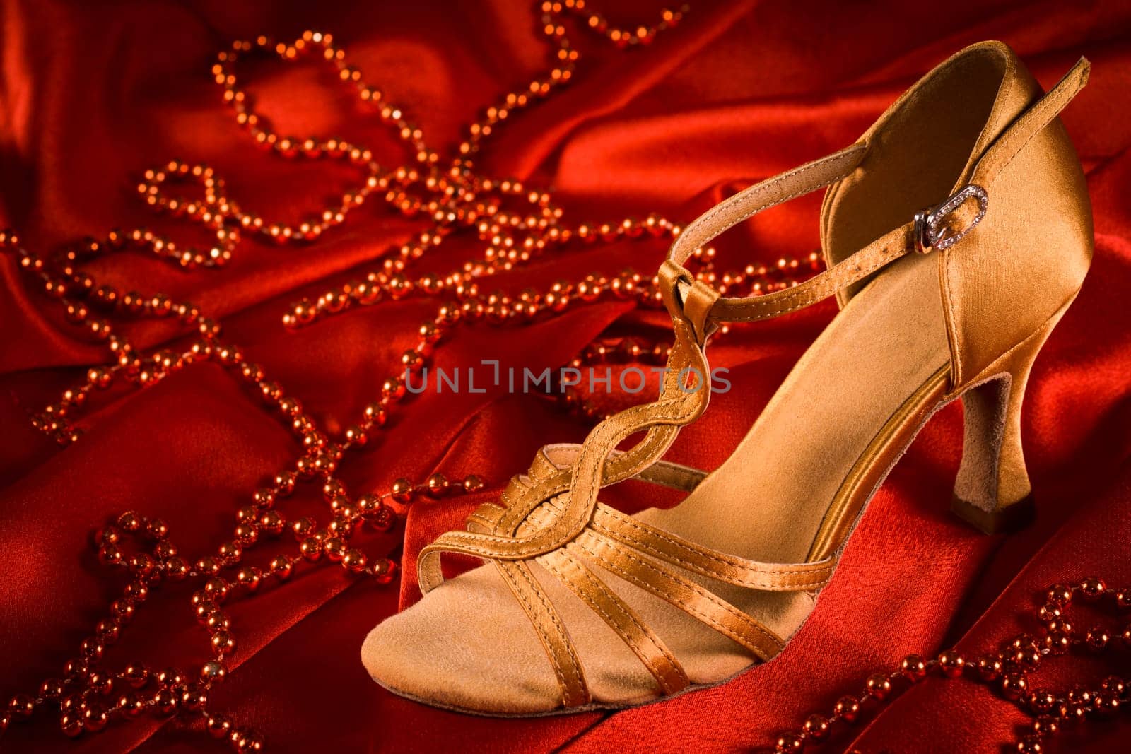 Golden latin dance shoe is on red satin background with red pearls.