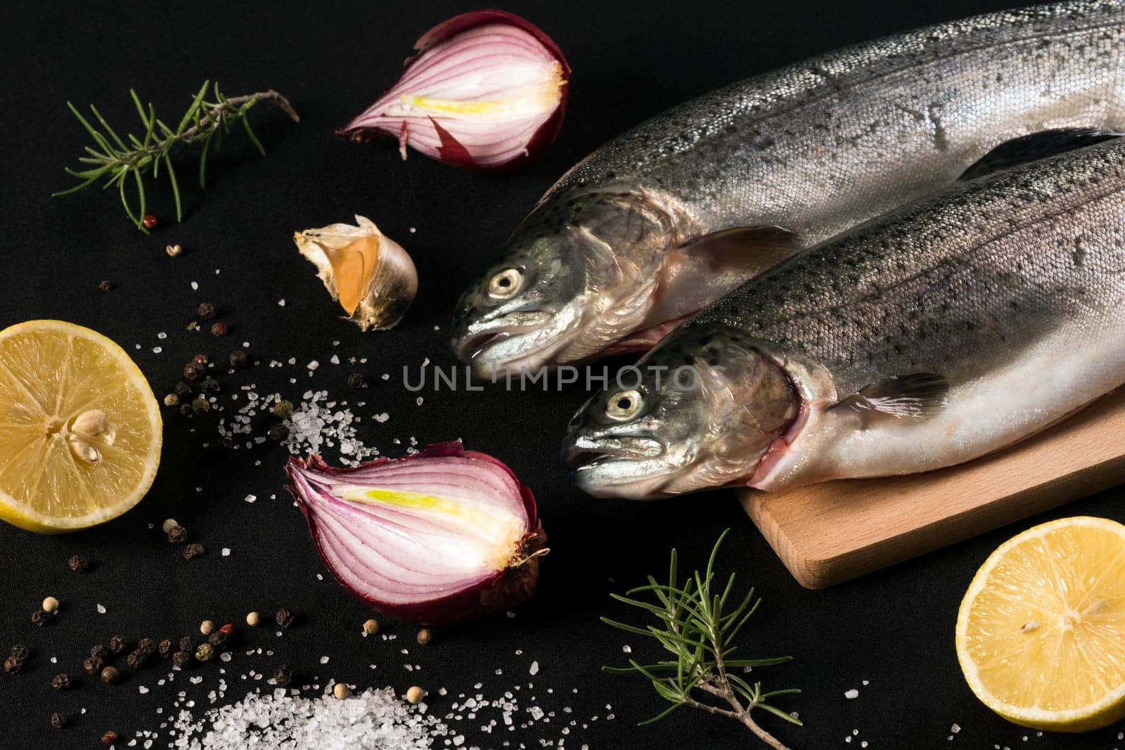 Raw trout fish with salt, pepper, lemon, garlic, onion and rosemary on black background.