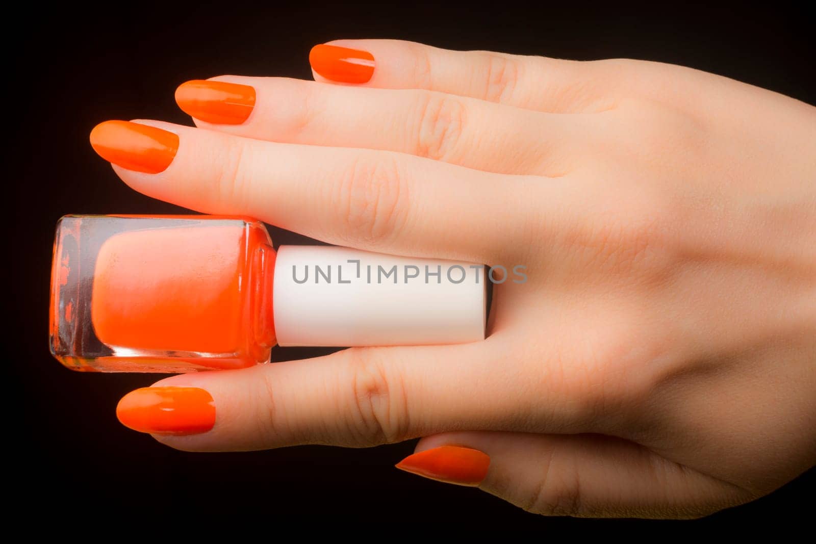 Female hand with fingers holds orange nail polish on black background, orange colored manicure.