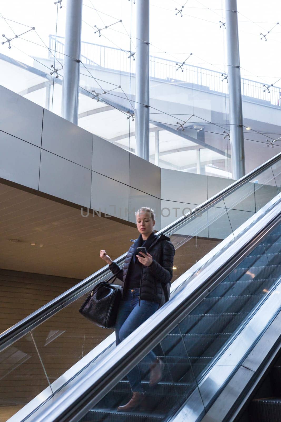 Young businesswoman with large black bag and mobile phone getting down on the escalator during business trip in modern city.
