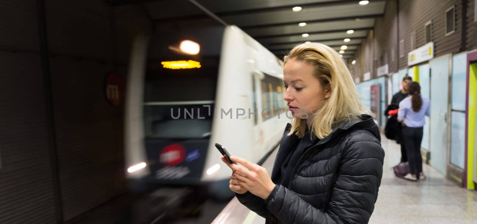 Young casual woman using mobile phone in her hand waiting on the platform of a metro station for metro to arrive. Public transport.
