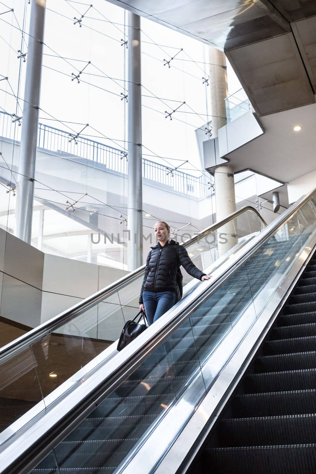 Young businesswoman with large black bag and mobile phone getting down on the escalator during business trip in modern city.