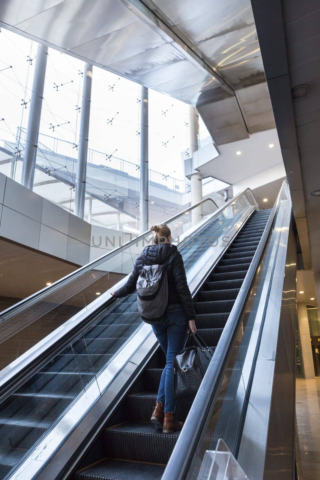 Young businesswoman with large black bag and mobile phone getting up the escalator during business trip in modern city.