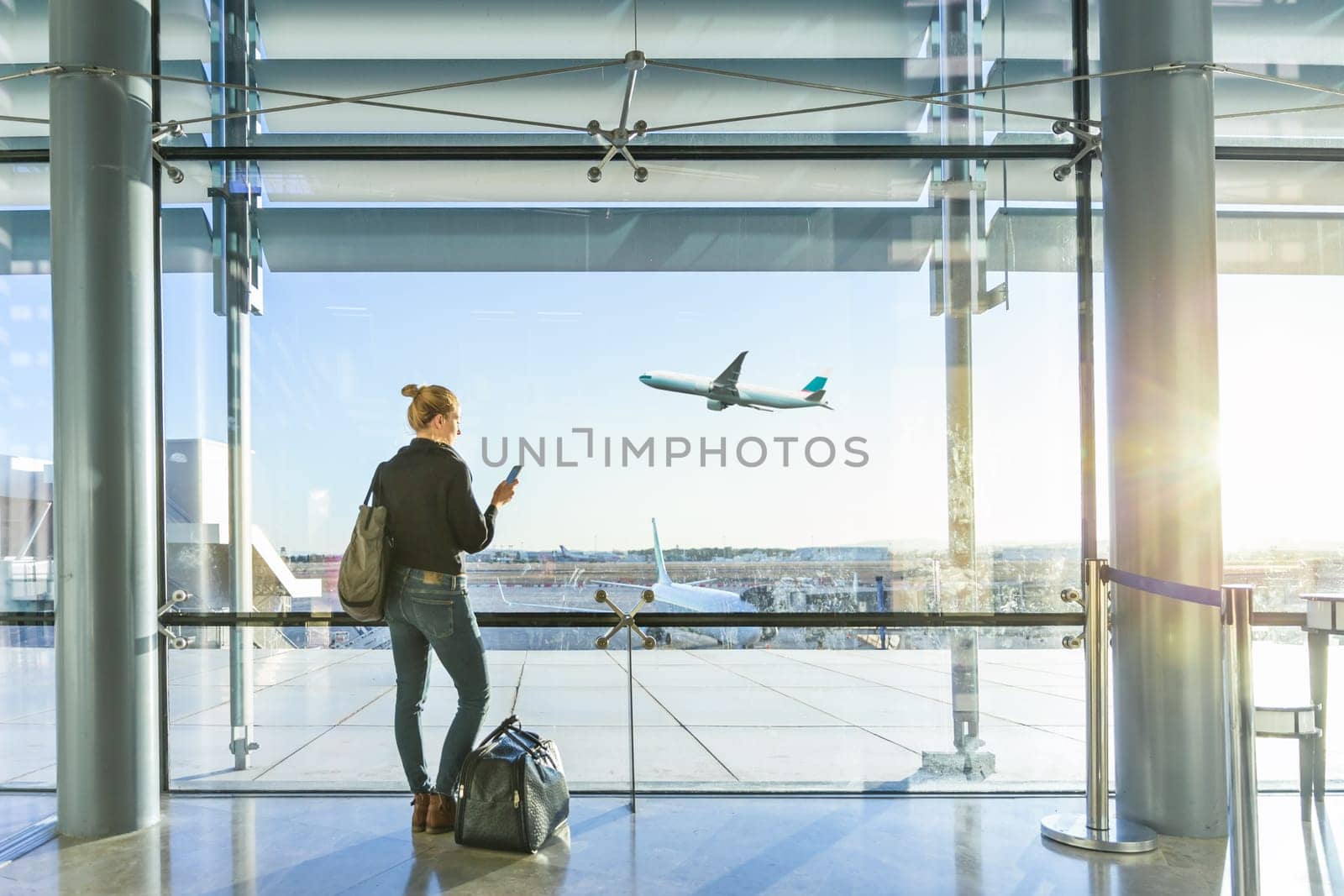 Young casual female traveler at airport, holding smart phone device, looking through the airport gate windows at planes on airport runway.