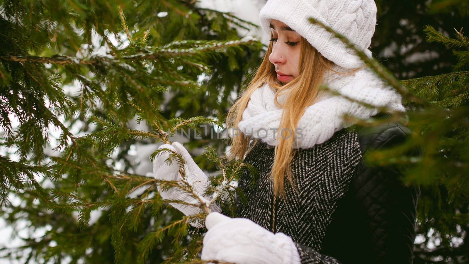 young woman in a forest with fir and pine trees on the street, hands touch the branches of trees, considering them. peridium winter, warm clothes, knitted mittens and scarf white