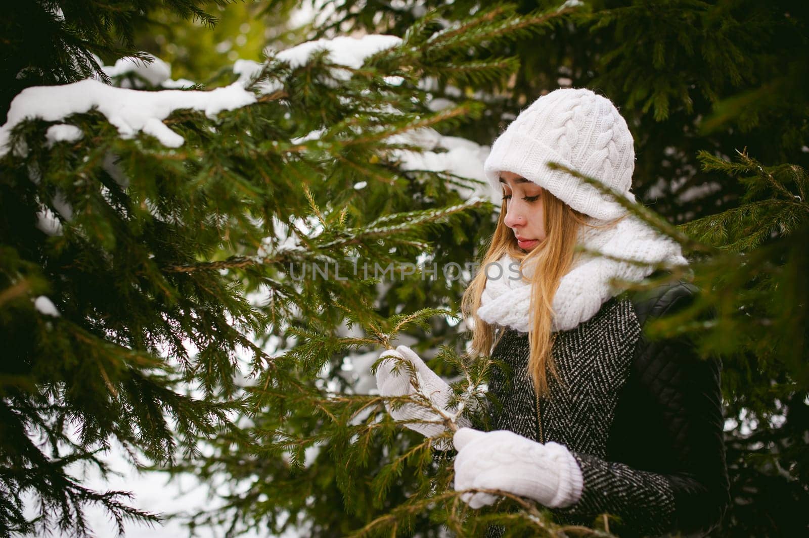 young woman in a forest with fir and pine trees on the street, hands touch the branches of trees, considering them. peridium winter, warm clothes, knitted mittens and scarf white