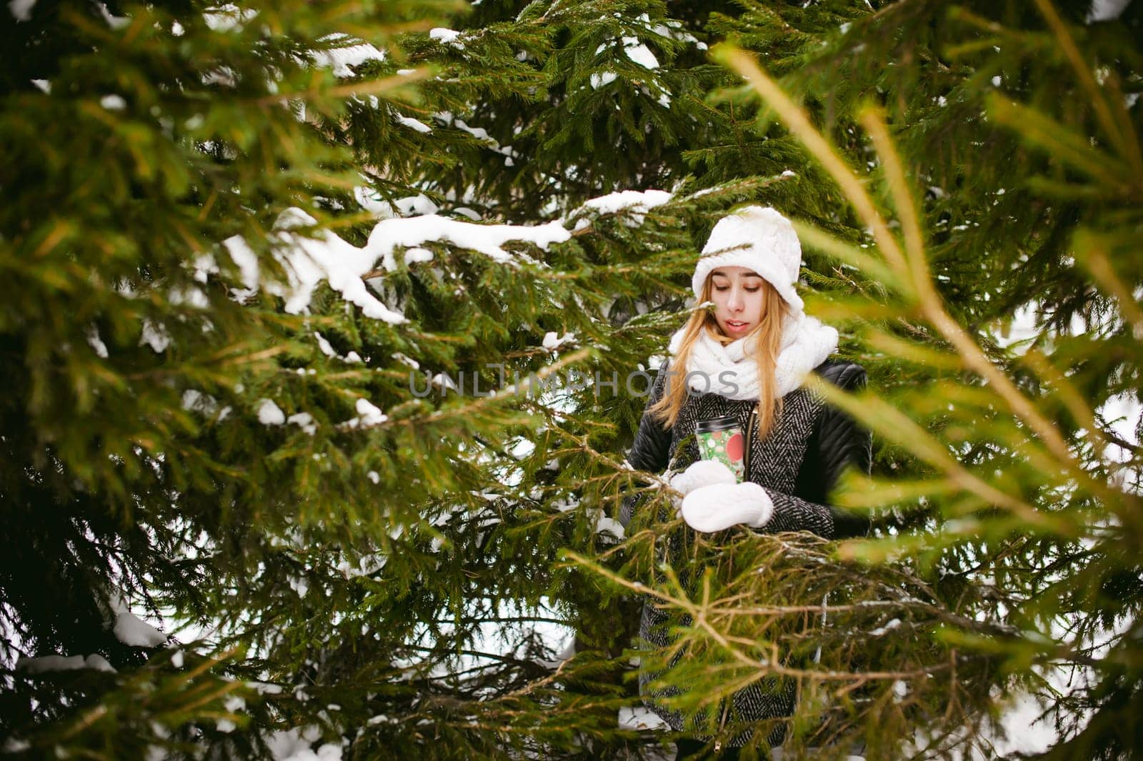 Portrait of cute woman in white scarf and hat knitted coat on outdoors background of snow and blurred fir branches in winter. girl holds a coffee cup in hand, christmas takeaway coffee