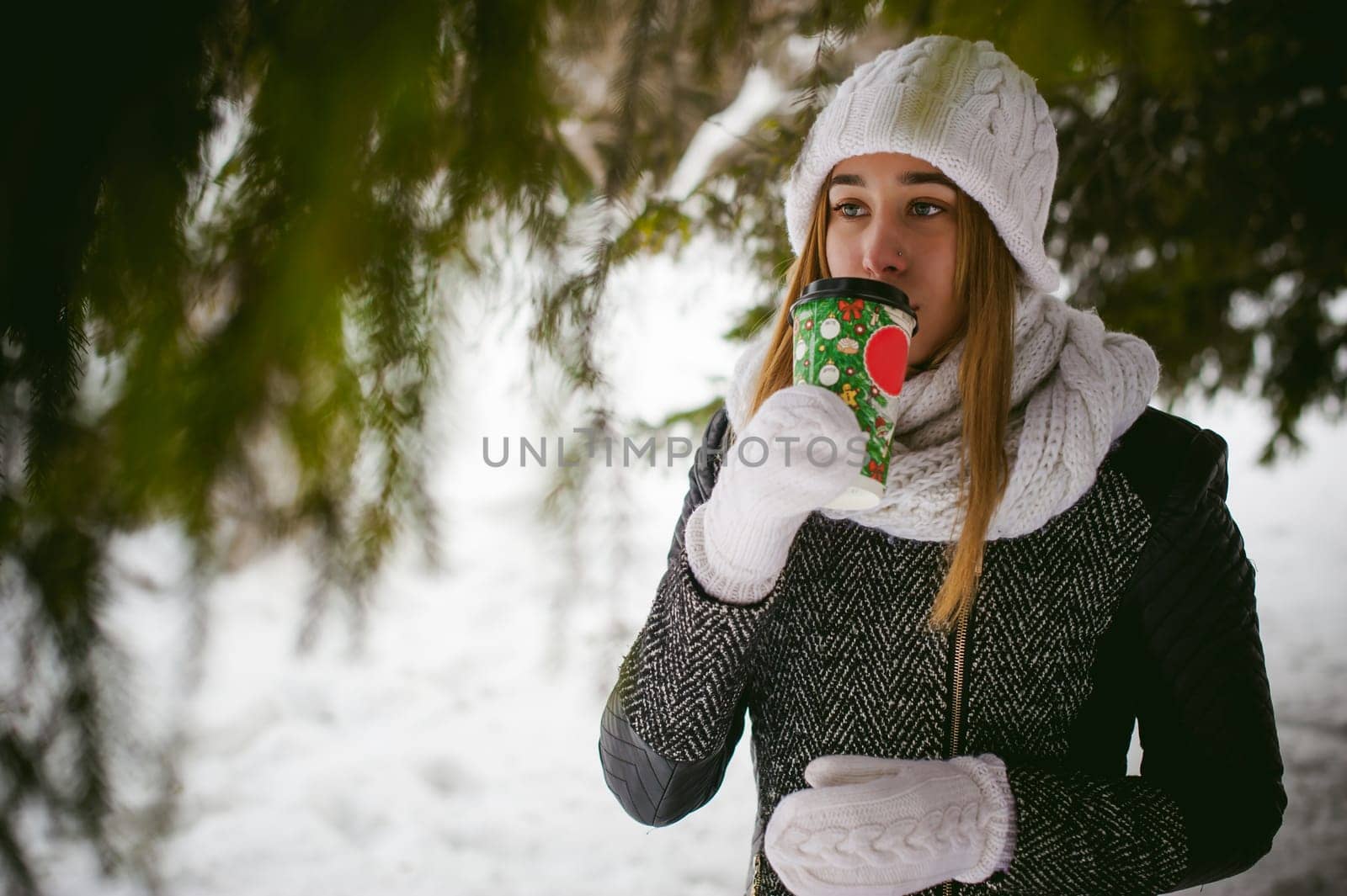 Portrait of cute woman in white scarf and hat knitted coat on outdoors background of snow and blurred fir branches in winter. girl holds a coffee cup in hand, christmas takeaway coffee