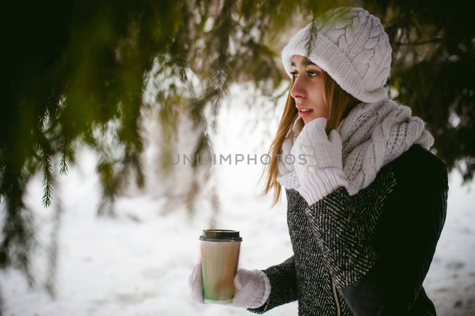 Portrait of cute woman in white scarf and hat knitted coat on outdoors background of snow and blurred fir branches in winter. girl holds a coffee cup in hand, christmas takeaway coffee