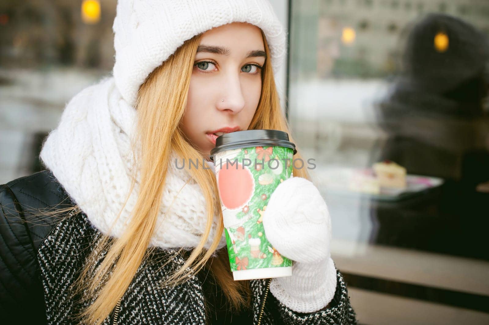 woman in white scarf and hat drinks coffee. cute young blonde girl sitting on a bench near the coffee shop, drinking takeaway coffee, holding Christmas paper cup in his hands. looking at the camera